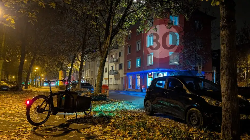 Eine ruhige, von Bäumen gesäumte Straße bei Nacht, bedeckt mit Herbstlaub. Ein Lastenfahrrad ist auf dem Gehweg geparkt, und ein kleines schwarzes Auto steht am Straßenrand. Die Gebäude sind beleuchtet, und ein Tempo-30-Schild ist zu sehen.