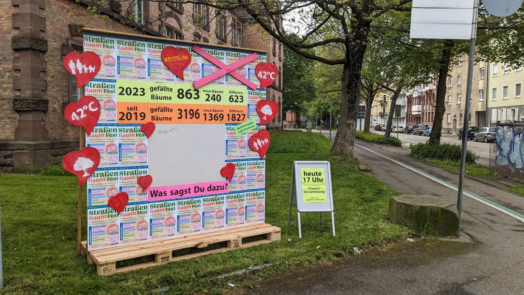 Eine bunte Schautafel auf dem Bürgersteig zeigt Statistiken über Bäume, mit roten Herzen und Zahlen und deutschem Text. Ein gelbes Schild in der Nähe kündigt eine Veranstaltung um 17 Uhr an. Die Straße im Hintergrund ist von Bäumen und Gebäuden gesäumt.