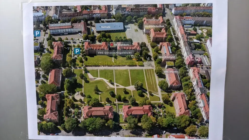 Luftaufnahme eines Parks, der von Gebäuden umgeben ist, mit beschrifteten Standorten wie Parkplätzen, einer Bibliothek, Klassenzimmern und Schulen. Wege, Bäume und Grünflächen sind im gesamten Gebiet zu sehen.