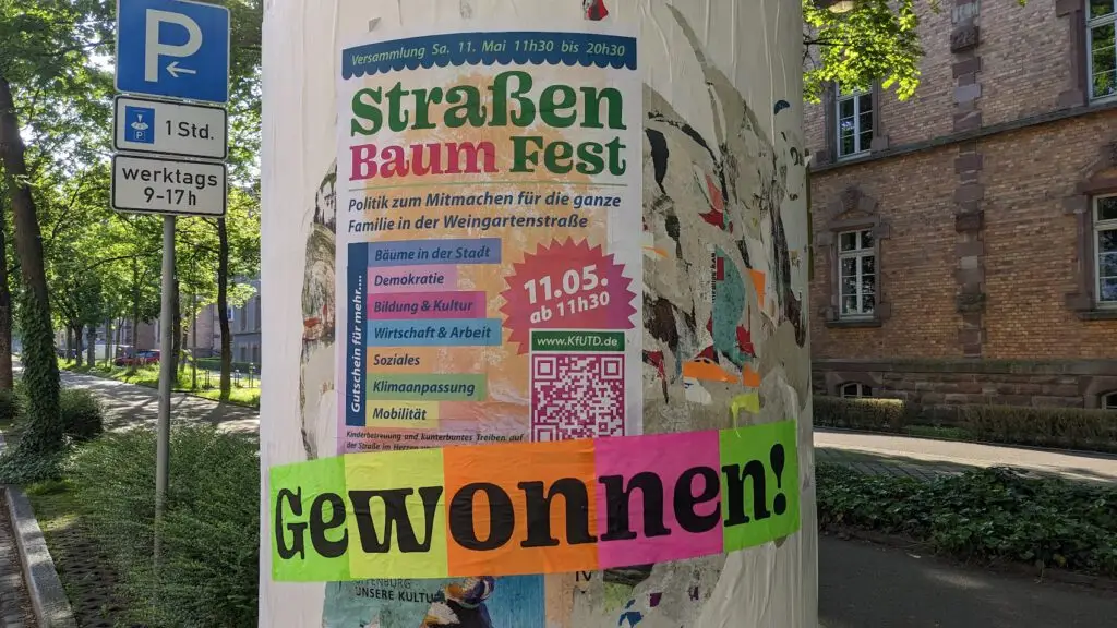 Ein buntes Plakat an einem Pfahl wirbt für das Straßen Baum Fest am 11. Mai mit Aktivitäten für Familien. Ein heller Streifen mit dem Wort Gewonnen! (Gewonnen!) ist am unteren Rand zu sehen.