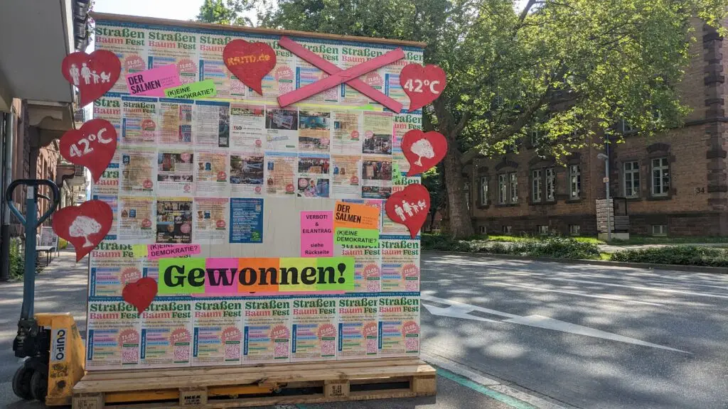 Eine Holztafel auf einer Palette ist mit bunten Plakaten, roten Herzausschnitten mit der Aufschrift 42°C und einem großen Schild mit der Aufschrift Gewonnen! auf einer Stadtstraße mit Bäumen und historischen Gebäuden im Hintergrund bedeckt.