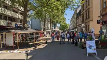Eine belebte Straßenszene zeigt Menschen, die auf einem Markt unter Bäumen spazieren gehen und sich versammeln, mit Verkaufsständen, Bänken und Gebäuden auf beiden Seiten der sonnigen Straße.