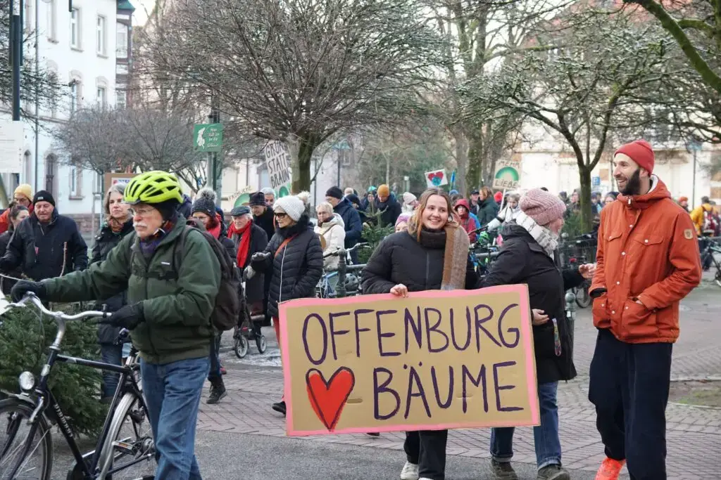 Eine Gruppe von Menschen nimmt an einem Protest im Freien teil; zwei lächelnde Frauen halten ein großes Schild mit der Aufschrift Offenburg ♥ Bäume, während andere in der Nähe auf einer Stadtstraße spazieren gehen oder Fahrrad fahren.
