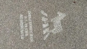 Weiße Kreidezeichnungen auf dem Pflaster zeigen eine vertikale Linie aus Kreisen und Rechtecken auf der linken Seite und ein stilisiertes geflügeltes Tier, möglicherweise ein Pferd oder einen Pegasus, auf der rechten Seite. Die Oberfläche ist rauer grauer Asphalt.