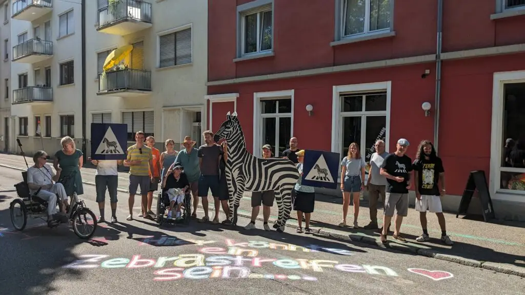 Eine Gruppe von Menschen steht auf einer sonnigen Straße und hält Plakate mit Fußgängerübergangssymbolen und einem großen Zebra-Ausschnitt in der Hand. Auf der Straße steht mit Kreide geschrieben "Ich könnte ein Zebrastreifen sein..." auf Deutsch.