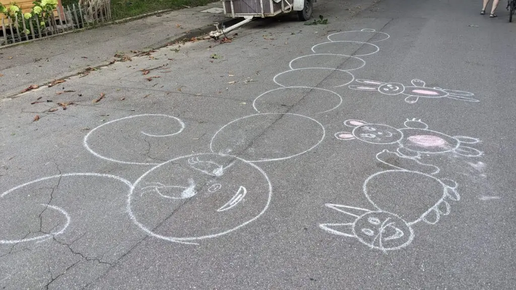 Bunte Kreidezeichnungen auf dem Bürgersteig zeigen eine große Raupe mit einem lächelnden Gesicht, ein Kaninchen, ein Schwein und eine Katze. Im Hintergrund sind ein Zaun, ein Wagen und Gras zu sehen.