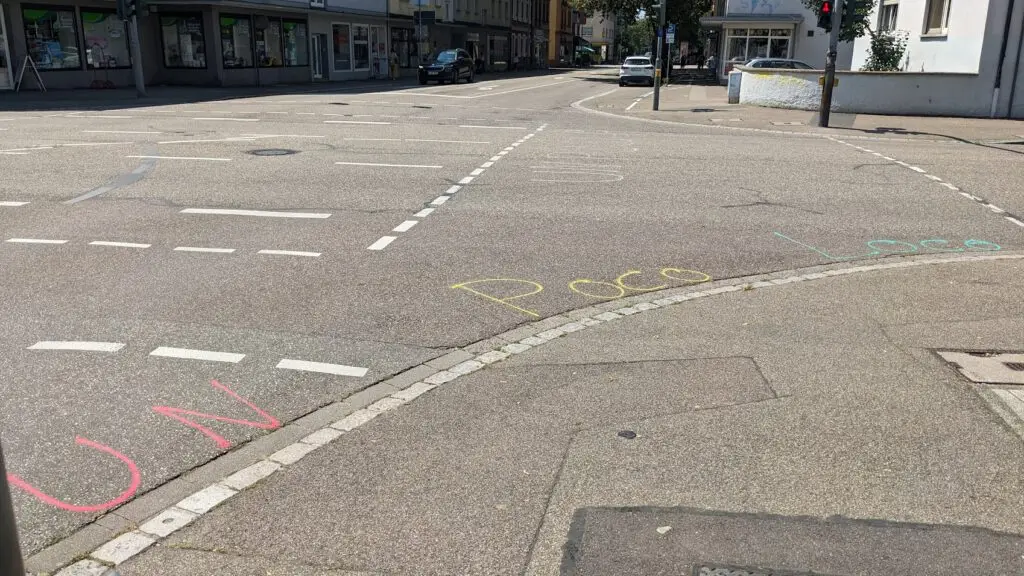Eine Straßenecke mit den bunten Kreideschriftzügen "UN", "Poco" und "Loco" auf dem Bürgersteig. Die Kreuzung ist leer, mit Gebäuden und Geschäften im Hintergrund.