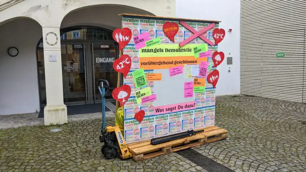Eine bunte Protesttafel auf einer Palette, bedeckt mit Plakaten und Schildern über Demokratie, Klimawandel und Straßenproteste, steht vor einem Hauseingang an einer Kopfsteinpflasterstraße. Rote Herzen und 42°C-Schilder sind angebracht.