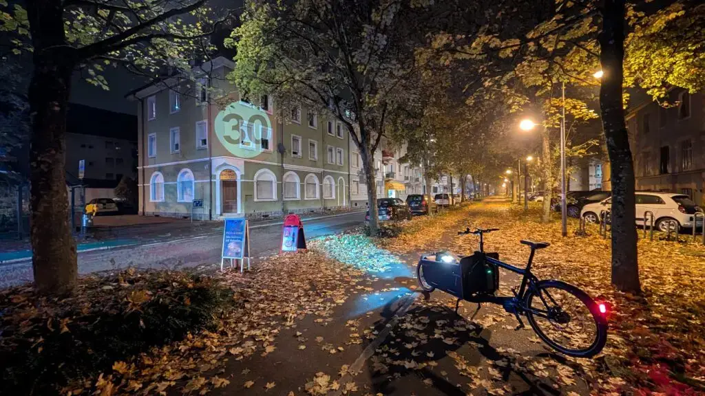 Ein Fahrrad mit einem hellen Scheinwerfer projiziert ein Tempo-30-Schild auf eine mit Herbstlaub bedeckte Straße bei Nacht. Straßenlaternen beleuchten parkende Autos und Wohnhäuser im Hintergrund.
