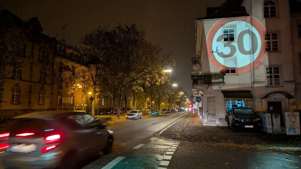 Eine nächtliche Straßenszene mit einem vorbeifahrenden Auto und einem großen beleuchteten Tempo 30-Schild, das auf die Seite eines Gebäudes projiziert wird. Straßenlaternen und geparkte Autos säumen die nasse Straße.