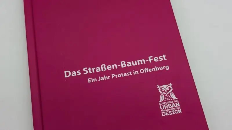 Ein rosa Buch mit dem Titel Das Straßen-Baum-Fest: Ein Jahr Protest in Offenburg, mit weißem Text und einem Eulenlogo für Urban Design. Das Buch ist als Teil einer Reihe über Urban Transformation Design gekennzeichnet.