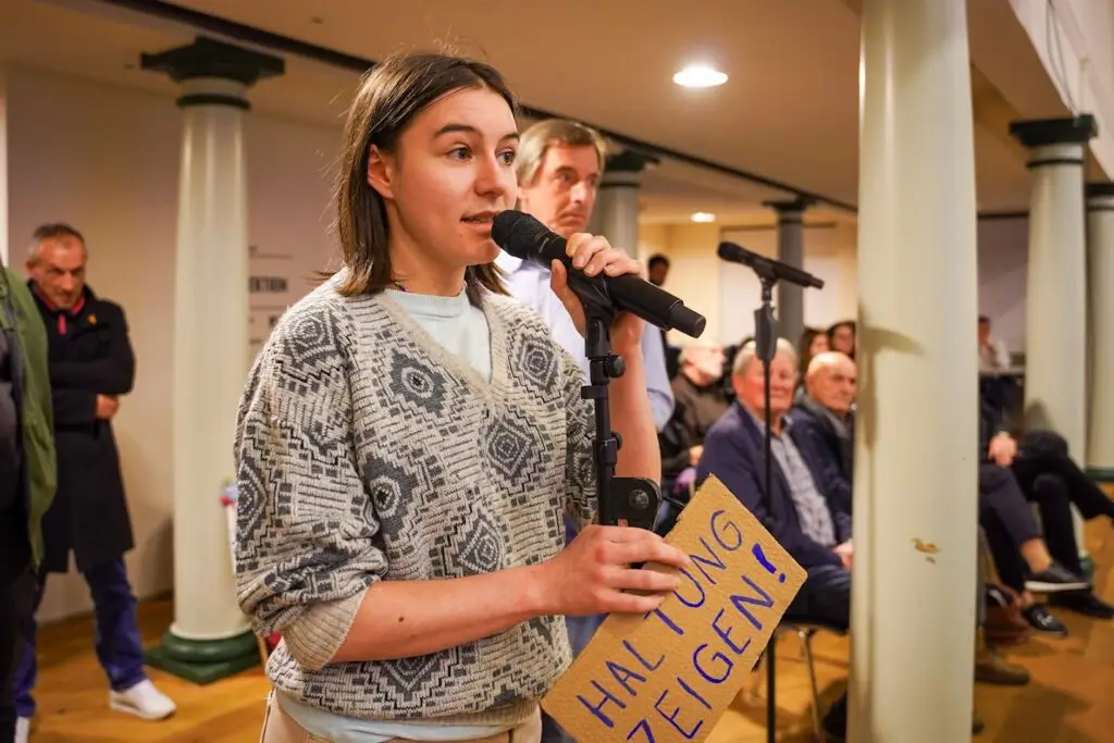 Eine Frau spricht bei einer Indoor-Veranstaltung in ein Mikrofon und hält ein Pappschild mit der Aufschrift "HALTUNG ZEIGEN!" in die Höhe. Im Hintergrund sitzen und stehen Menschen und hören aufmerksam zu.