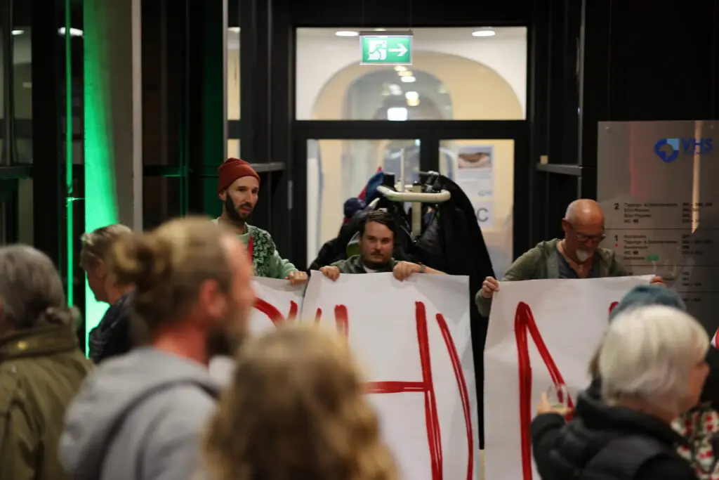 Eine Gruppe von Menschen in einem Haus hält große weiße Schilder mit roten Buchstaben. Drei Personen stehen vorne mit den Schildern, während andere zusehen oder Fotos machen. Hinter den Schilderhaltern hängen Mäntel an einem Gestell.