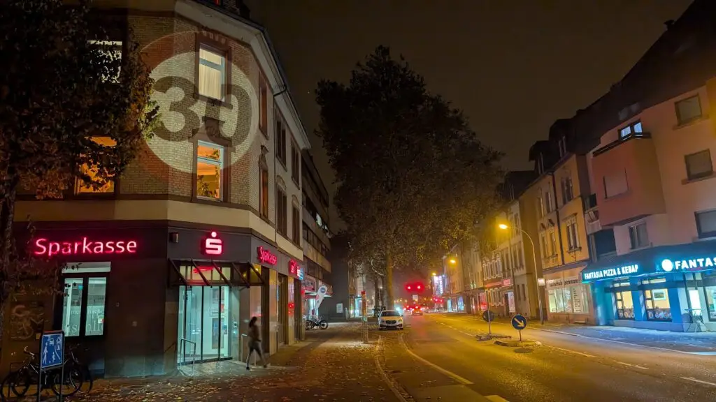 Eine nächtliche Stadtstraße mit beleuchteten Ladenschildern, darunter die Sparkasse. Eine Person verlässt das Gebäude, und entlang der Straße sind Autos geparkt. Bäume säumen die Straße, und eine große 39 ist auf das Eckgebäude projiziert.