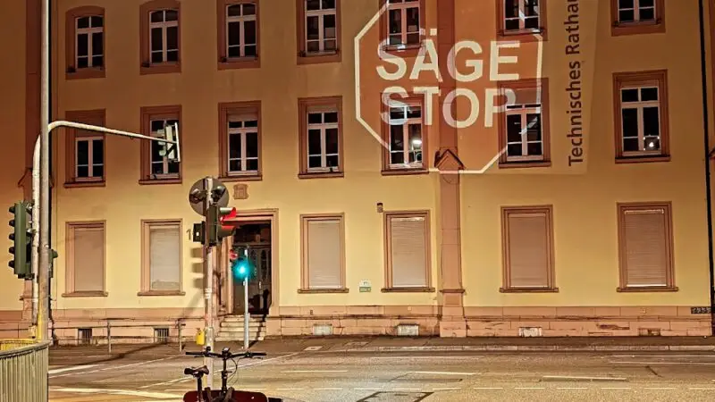 Ein Lastenfahrrad mit rotem Rücklicht wartet an einer nächtlichen Ampel vor dem Technischen Rathaus, das "SAGE STOP"-Schild ist zu sehen - eine von urbanem Leben und Stadtklima geprägte Szene, auch wenn keine Bäume das verschlossene Gebäude säumen.