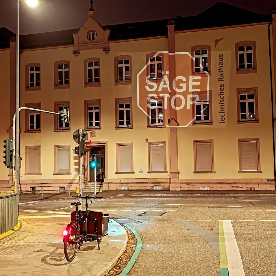 Ein Lastenfahrrad mit rotem Rücklicht wartet an einer nächtlichen Ampel vor dem Technischen Rathaus, das "SAGE STOP"-Schild ist zu sehen - eine von urbanem Leben und Stadtklima geprägte Szene, auch wenn keine Bäume das verschlossene Gebäude säumen.