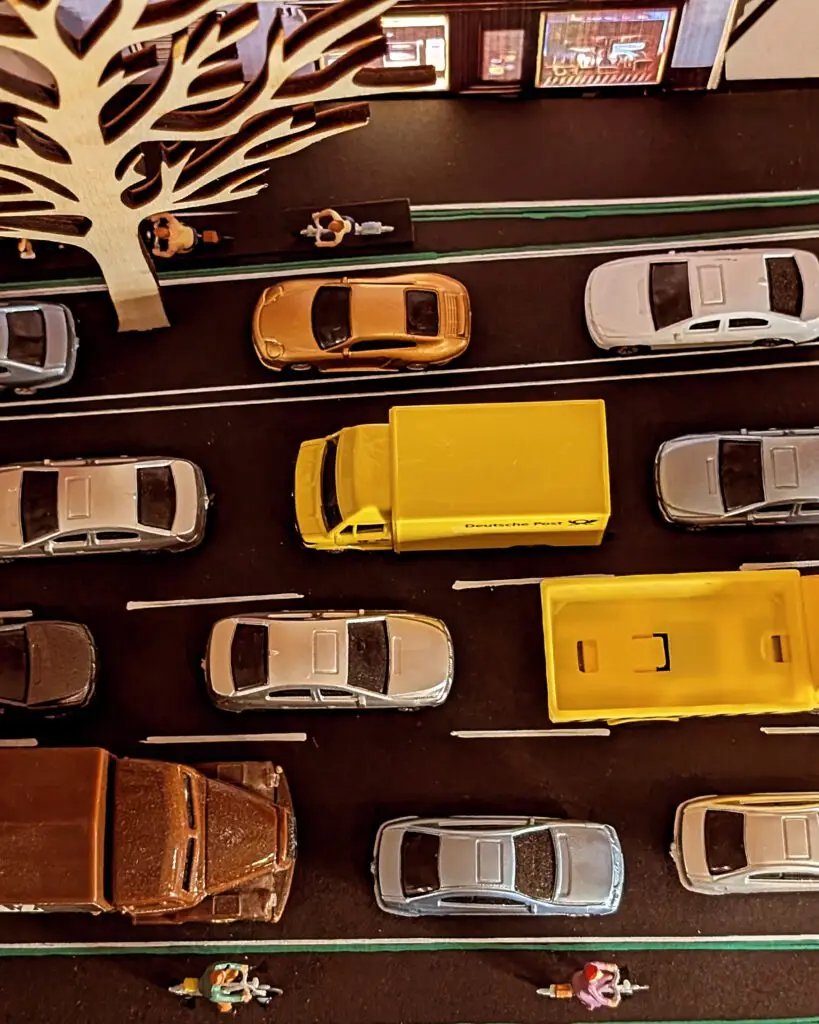 Eine Draufsicht auf eine Spielzeugstadt mit mehreren Spielzeugautos, zwei gelben Lieferwagen, einem Baum, winzigen Fußgängerfiguren und einer Reihe von Gebäuden am oberen Bildrand.