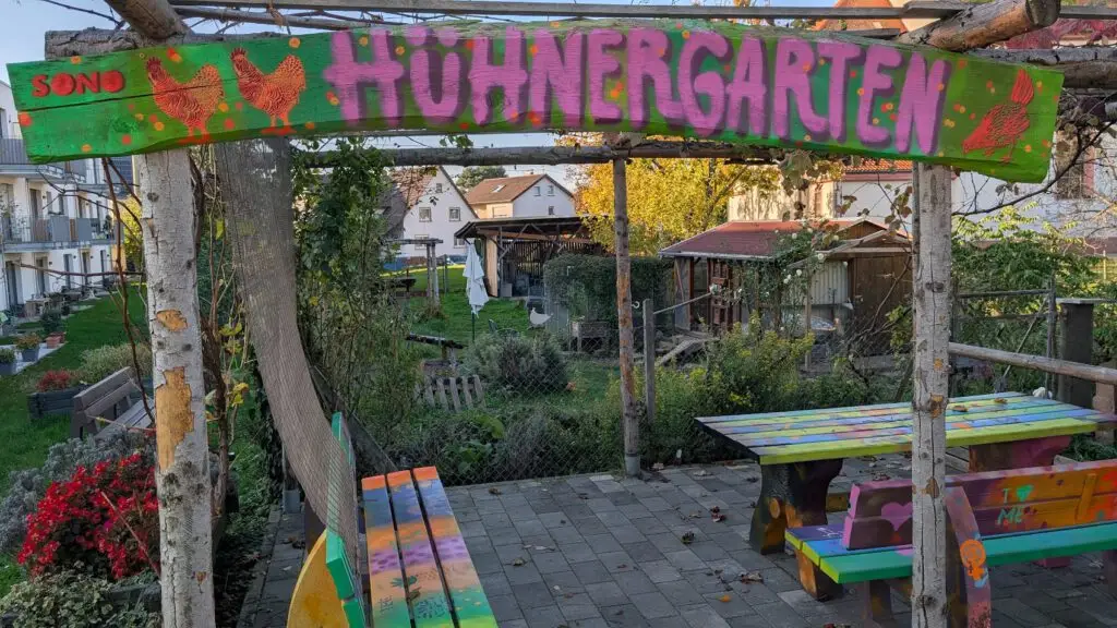 Ein buntes Schild mit der Aufschrift HÜHNERGARTEN hängt über einer Terrasse mit bemalten Bänken und Tischen, die einen eingezäunten Garten mit Grünpflanzen und kleinen Strukturen in einem Wohnviertel überblickt.