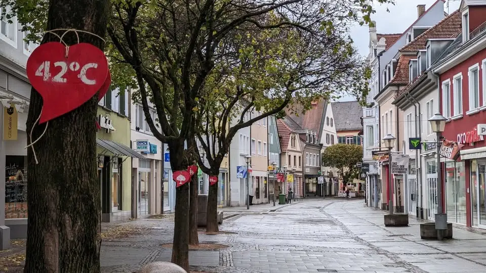 Eine verlassene, von Bäumen gesäumte Straße in einer europäischen Stadt mit roten herzförmigen Schildern, die an den Bäumen hängen, eines mit der Aufschrift 42°C. Die Geschäfte entlang der Straße scheinen geschlossen und das Wetter ist bedeckt.