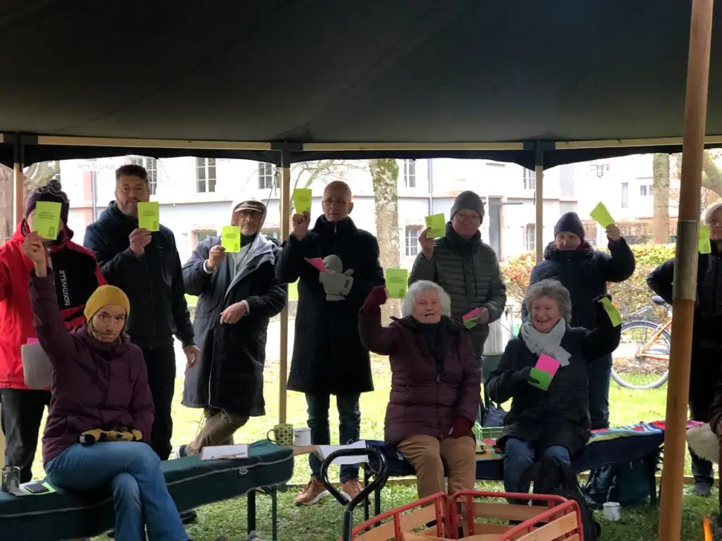Eine Gruppe von warm gekleideten Menschen sitzt und steht unter einem Vordach im Freien, jeder hält eine grüne oder gelbe Karte hoch und nimmt an einer Gruppenaktivität oder Abstimmung teil. Im Hintergrund sind Bäume und Gebäude zu sehen.