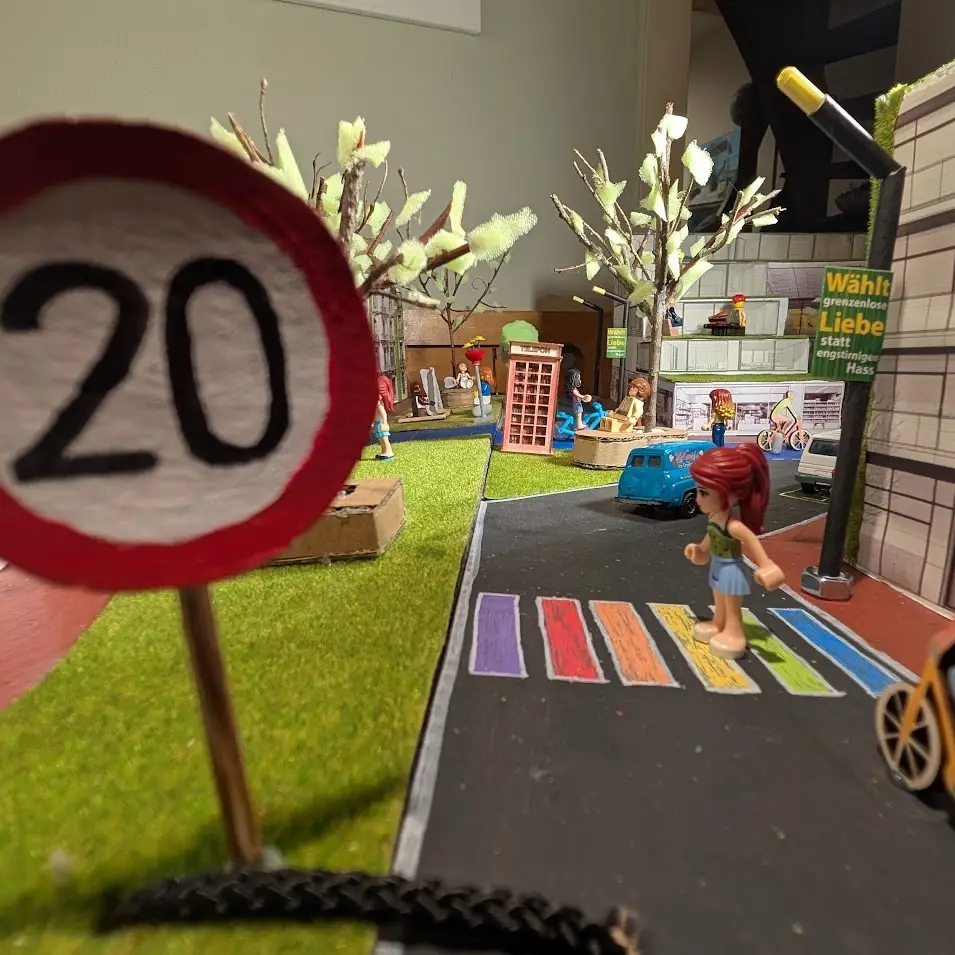 Ein Diorama mit Spielzeugfiguren zeigt ein Schild mit einer Geschwindigkeitsbegrenzung von 20 km/h, einen regenbogenfarbenen Zebrastreifen, Bäume, einen blauen Lieferwagen, Fahrräder und verschiedene Figuren, darunter eine, die die Straße überquert, alles in einer Miniaturstadtszene.