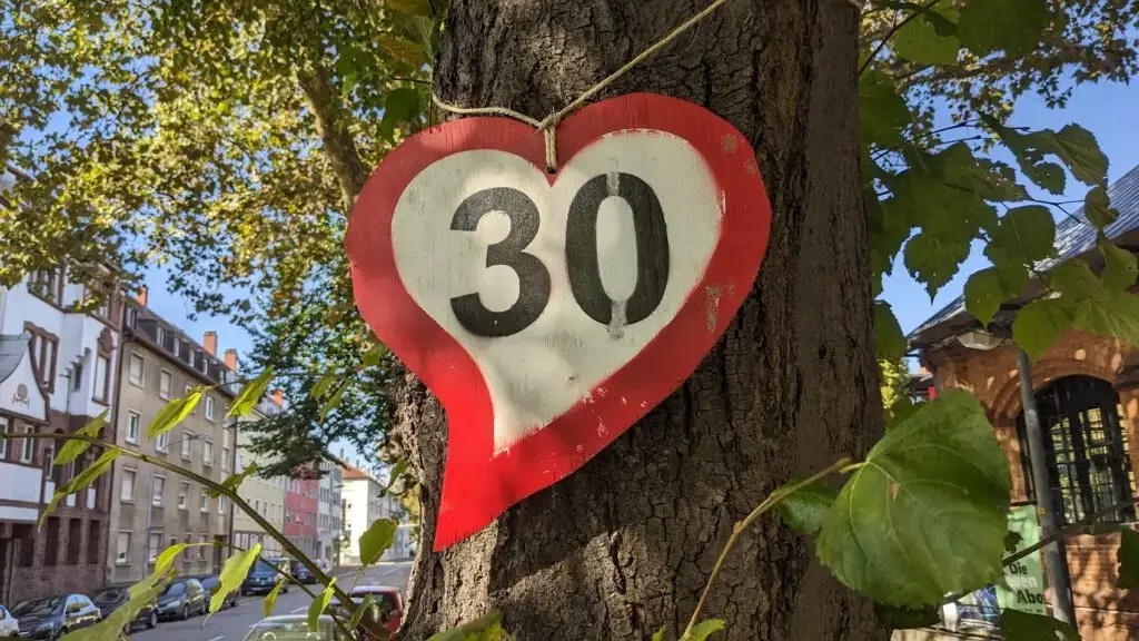 Ein rot-weißes herzförmiges Schild mit der Zahl 30 hängt an einem Baumstamm, im Hintergrund sind grüne Blätter und Gebäude an einem sonnigen Tag zu sehen.