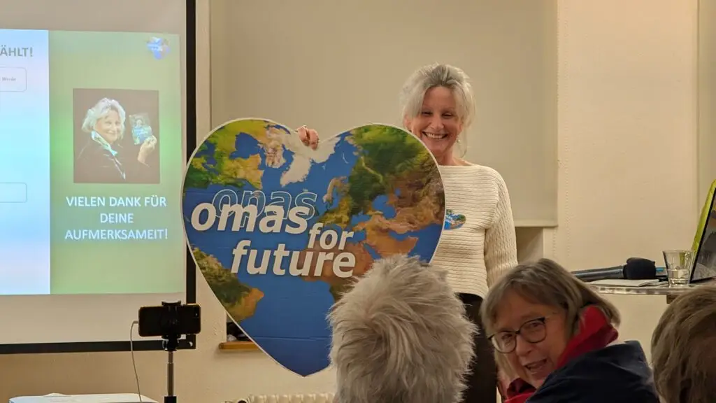 Eine lächelnde Frau hält ein großes, herzförmiges Schild mit der Aufschrift omas for future und einer Europakarte in der Hand. Auf einem Bildschirm im Hintergrund sind ihr Foto und ein deutscher Text zu sehen. Mehrere ältere Erwachsene sitzen da und schauen zu.