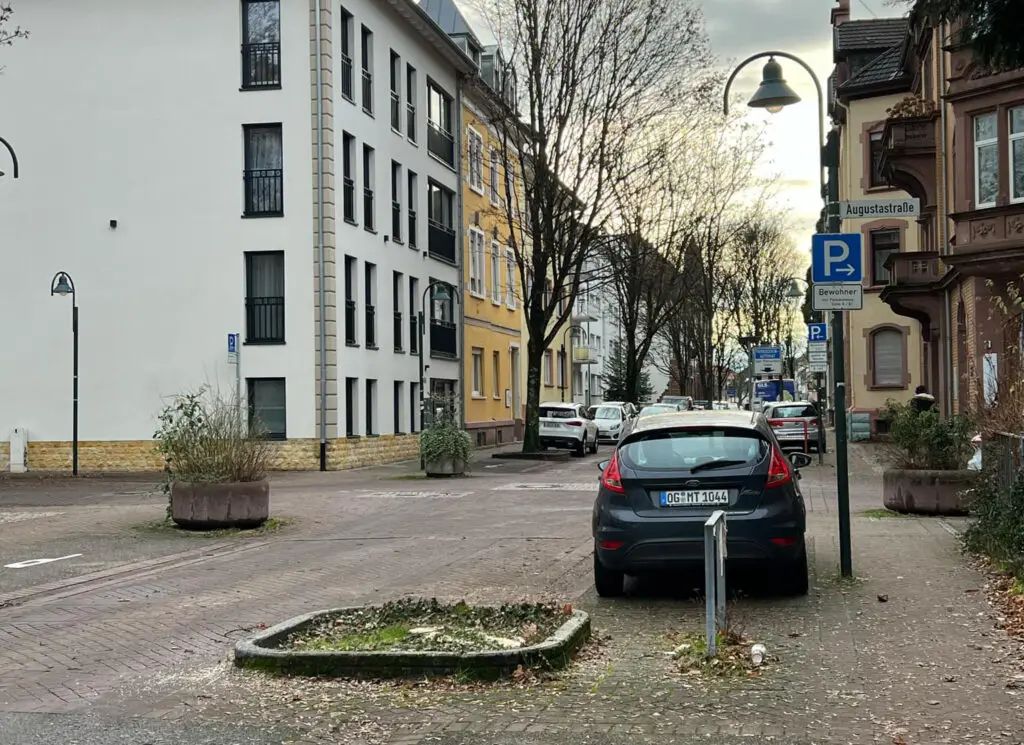 Ein geparktes schwarzes Auto neben einem Bewohnerparken-Schild in einer ruhigen städtischen Straße, die an einem bewölkten Tag von Gebäuden, Bäumen ohne Blätter und geparkten Fahrzeugen gesäumt ist.