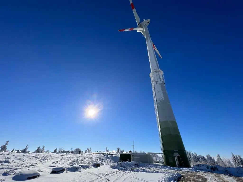 Eine große Windkraftanlage steht in einer schneebedeckten Landschaft bei strahlend blauem Himmel und Sonnenschein. Am Fuße des Windrads sind Bäume und ein kleines Gebäude zu sehen.