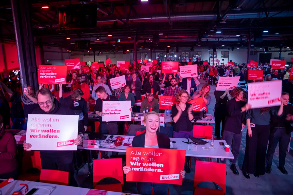 Eine große Gruppe von Menschen auf einer politischen Veranstaltung hält rote Schilder mit der Aufschrift "Alle wollen regieren. Wir wollen verändern." Der Raum ist hell mit rotem Licht beleuchtet, und viele Teilnehmer lächeln oder klatschen.