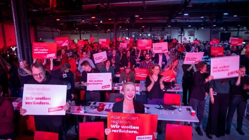 Eine große Gruppe von Menschen auf einer politischen Veranstaltung hält rote Schilder mit der Aufschrift "Alle wollen regieren. Wir wollen verändern." Der Raum ist hell mit rotem Licht beleuchtet, und viele Teilnehmer lächeln oder klatschen.