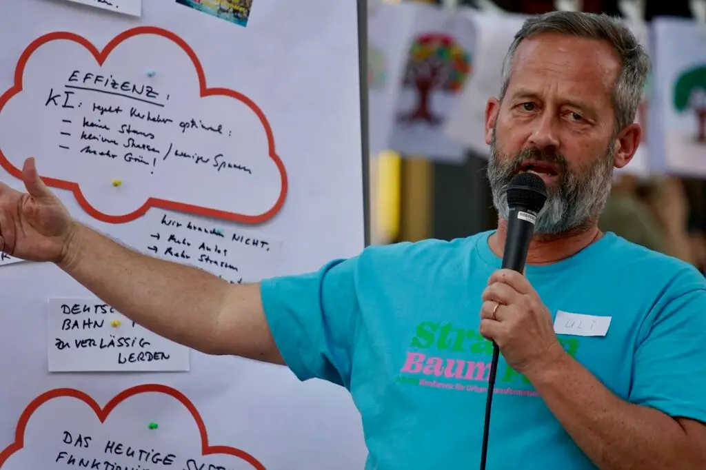 Ein Mann mit Bart und blauem Hemd spricht in ein Mikrofon und deutet auf ein großes Plakat mit handschriftlichen Notizen darauf. Das Plakat zeigt wolkenförmige Umrisse und ist im Freien aufgehängt.
