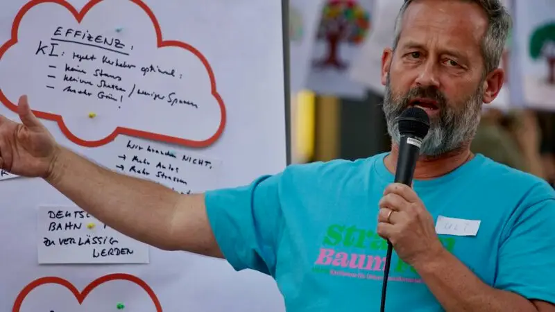 Ein Mann mit Bart und blauem Hemd spricht in ein Mikrofon und deutet auf ein großes Plakat mit handschriftlichen Notizen darauf. Das Plakat zeigt wolkenförmige Umrisse und ist im Freien aufgehängt.