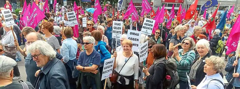 Eine große Gruppe von Menschen, darunter viele ältere Frauen, marschiert mit rosa Fahnen und Schildern mit der Aufschrift OMAS GEGEN RECHTS ("Großmütter gegen Rechts") bei einer Demonstration oder Kundgebung auf einer Straße in der Stadt.