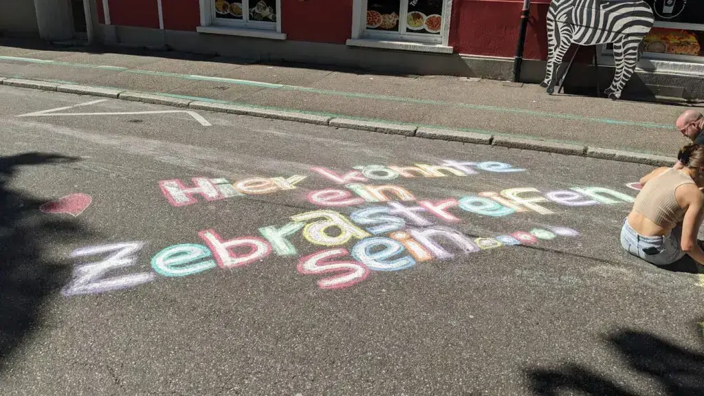 Bunte Kreideschrift auf einer Straße: "Hier könnte ein Zebrastreifen sein..." auf Deutsch. Zwei Menschen sitzen in der Nähe, und ein Teil einer Zebradekoration steht an einem Gebäude im Hintergrund.