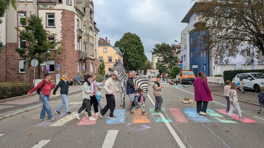 Eine Gruppe von Menschen, darunter Kinder und Erwachsene, überquert eine Straße auf einem mit bunter Kreide gezeichneten Zebrastreifen. Eine Person in einem Zebrakostüm geht mit ihnen. Gebäude und Bäume säumen die Straße an einem bewölkten Tag.