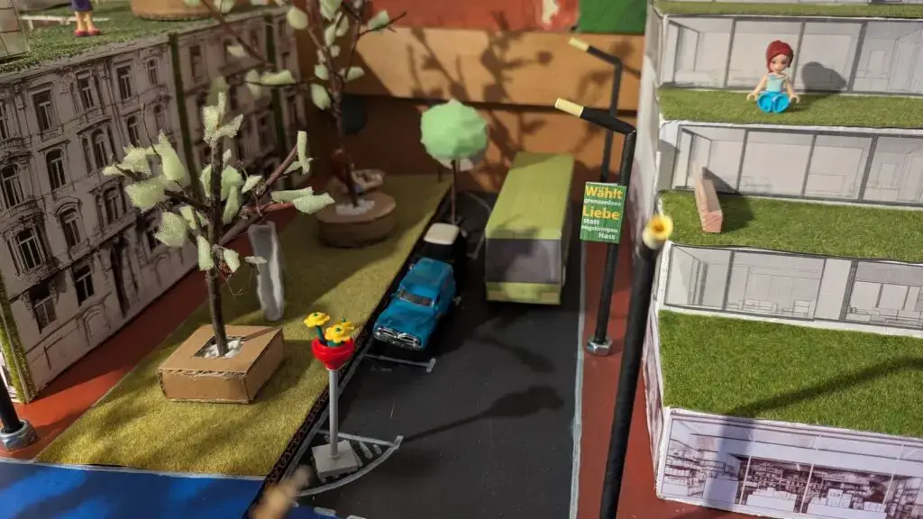 Eine Miniatur-Straßenszene mit Bäumen, Grasdächern, geparkten Autos und Papiergebäuden. Eine Figur sitzt auf einem Dach, und eine Straßenlaterne und eine Ampel sind in der Nähe eines blauen Autos und eines grünen Lastwagens zu sehen.