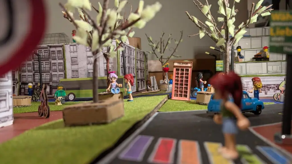 Eine bunte Miniaturstadtszene aus Papier und Spielzeugfiguren, mit Bäumen, Gebäuden, einer Telefonzelle, Fahrzeugen und einem Regenbogenüberweg im Vordergrund.