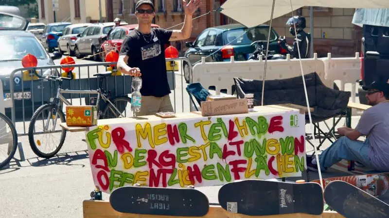 Ein Mann winkt und lächelt hinter einem Tisch im Freien, auf dem Skateboards und ein buntes Schild in deutscher Sprache stehen, das für die Beteiligung am öffentlichen Raum wirbt; eine weitere Person sitzt in der Nähe. Im Hintergrund sind Autos und Fahrräder geparkt.