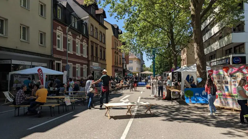 Ein lebhaftes Straßenfest mit Menschen, die an Tischen sitzen, spazieren gehen und Stände entlang einer sonnigen, von Bäumen gesäumten Straße besuchen, die von bunten Gebäuden und Zelten gesäumt ist.