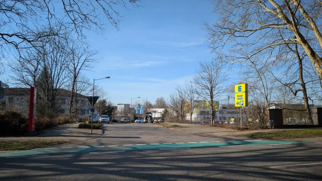 Eine ruhige Straße an einem sonnigen Tag, mit Bäumen auf beiden Seiten und ein paar geparkten Autos. Auf der rechten Seite sind mehrere Schilder für Edeka, einen deutschen Supermarkt, zu sehen. Der Himmel ist klar und blau.