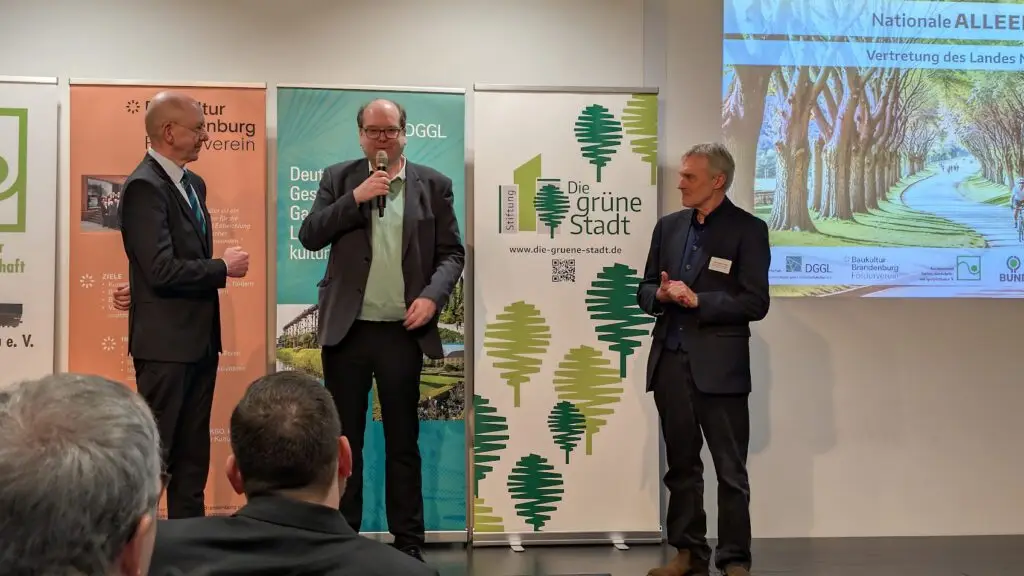 Drei Männer in Anzügen stehen auf einer Bühne und sprechen in Mikrofone vor Spruchbändern über grüne Städte und kulturelle Organisationen, während das Publikum sie beobachtet.