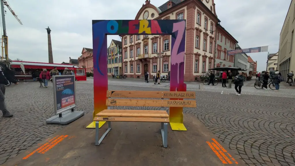 Eine bunte Bank mit der Aufschrift "Kein Platz für Rassismus!" steht auf einem Stadtplatz, umrahmt von einem Regenbogen. Im Hintergrund sind historische Gebäude und spazierende Menschen zu sehen.