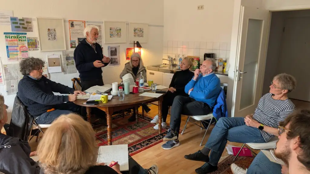 Eine Gruppe von Menschen sitzt in einem Kreis in einem gemütlichen Raum und diskutiert. Einige machen sich Notizen, während ein Mann steht und spricht. Papiere, Tassen und Dokumente sind auf dem Tisch ausgebreitet, und der Raum hat eine warme, informelle Atmosphäre.