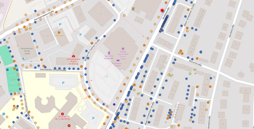 Eine detaillierte Straßenkarte mit Gebäuden, Parkplätzen, Straßen und Sehenswürdigkeiten, die mit farbigen Punkten in einem Stadtviertel markiert sind. Beschriftungen und Icons weisen auf Geschäfte, Parkplätze und öffentliche Einrichtungen hin.