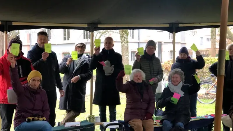 Eine Gruppe von Menschen, sowohl sitzend als auch stehend im Freien unter einem Vordach, hält grüne Karten hoch. Sie sind warm mit Mänteln und Mützen gekleidet, was auf kaltes Wetter hindeutet. Im Hintergrund sind Bäume und Fahrräder zu sehen.