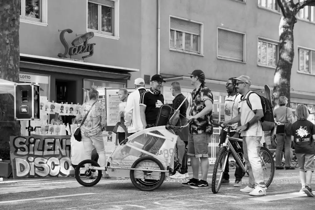 Eine Gruppe von Menschen steht draußen auf einer Stadtstraße in der Nähe einer Bar namens Saxy. Einige halten Fahrräder, einer schiebt ein Kind in einem Anhänger, und es sind Dekorationen und ein "Silent Disco"-Schild zu sehen. Das Foto ist schwarz-weiß.