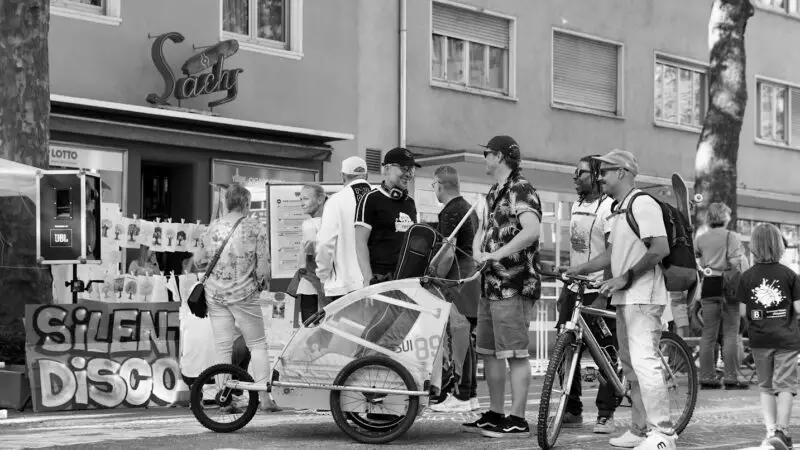 Eine Gruppe von Menschen steht draußen auf einer Stadtstraße in der Nähe einer Bar namens Saxy. Einige halten Fahrräder, einer schiebt ein Kind in einem Anhänger, und es sind Dekorationen und ein "Silent Disco"-Schild zu sehen. Das Foto ist schwarz-weiß.