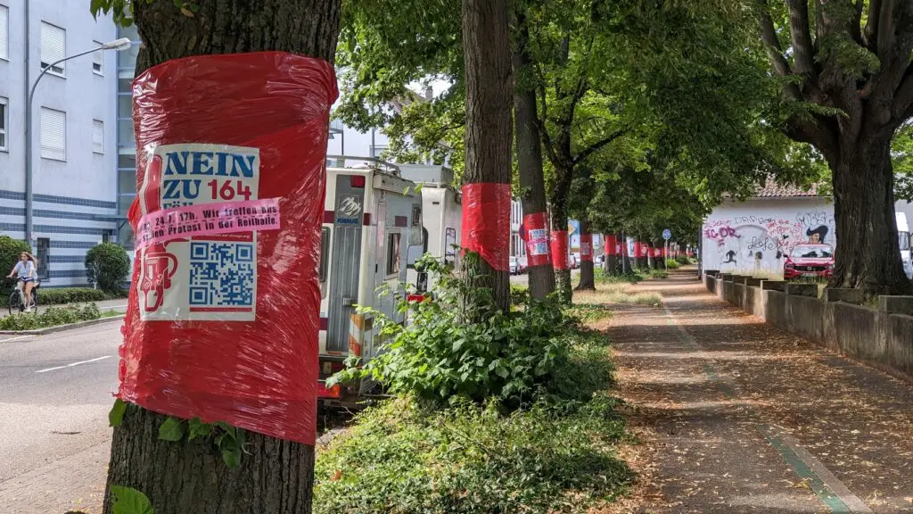 Bäume entlang eines Bürgersteigs sind mit rotem Plastik umwickelt und tragen Plakate mit der Aufschrift NEIN ZU 164 und einem QR-Code. Eine Person geht auf der Straße, und im Hintergrund ist ein Graffiti an einer Wand zu sehen.