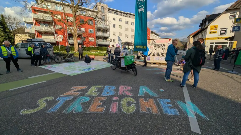 Menschen versammeln sich auf einer für den Verkehr gesperrten Stadtstraße, auf deren Bürgersteig mit bunter Kreide ZEBRA-STREICHEN geschrieben steht. Einige stehen in der Nähe von Fahrrädern und Informationsschildern, und im Hintergrund sind Gebäude zu sehen.
